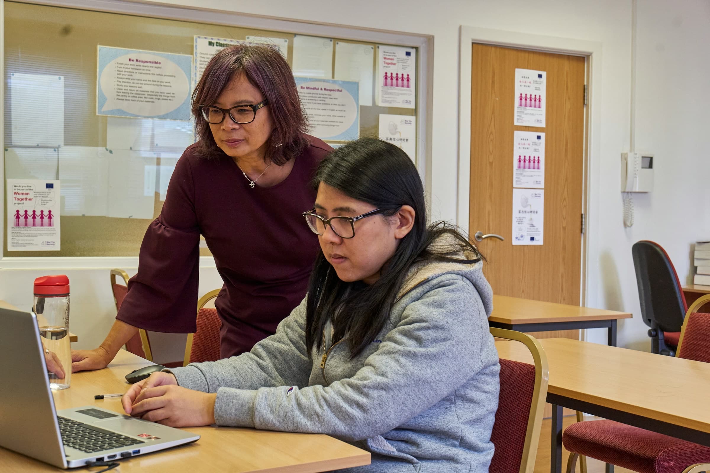 A tutor leaning in to help a student with her work on a laptop